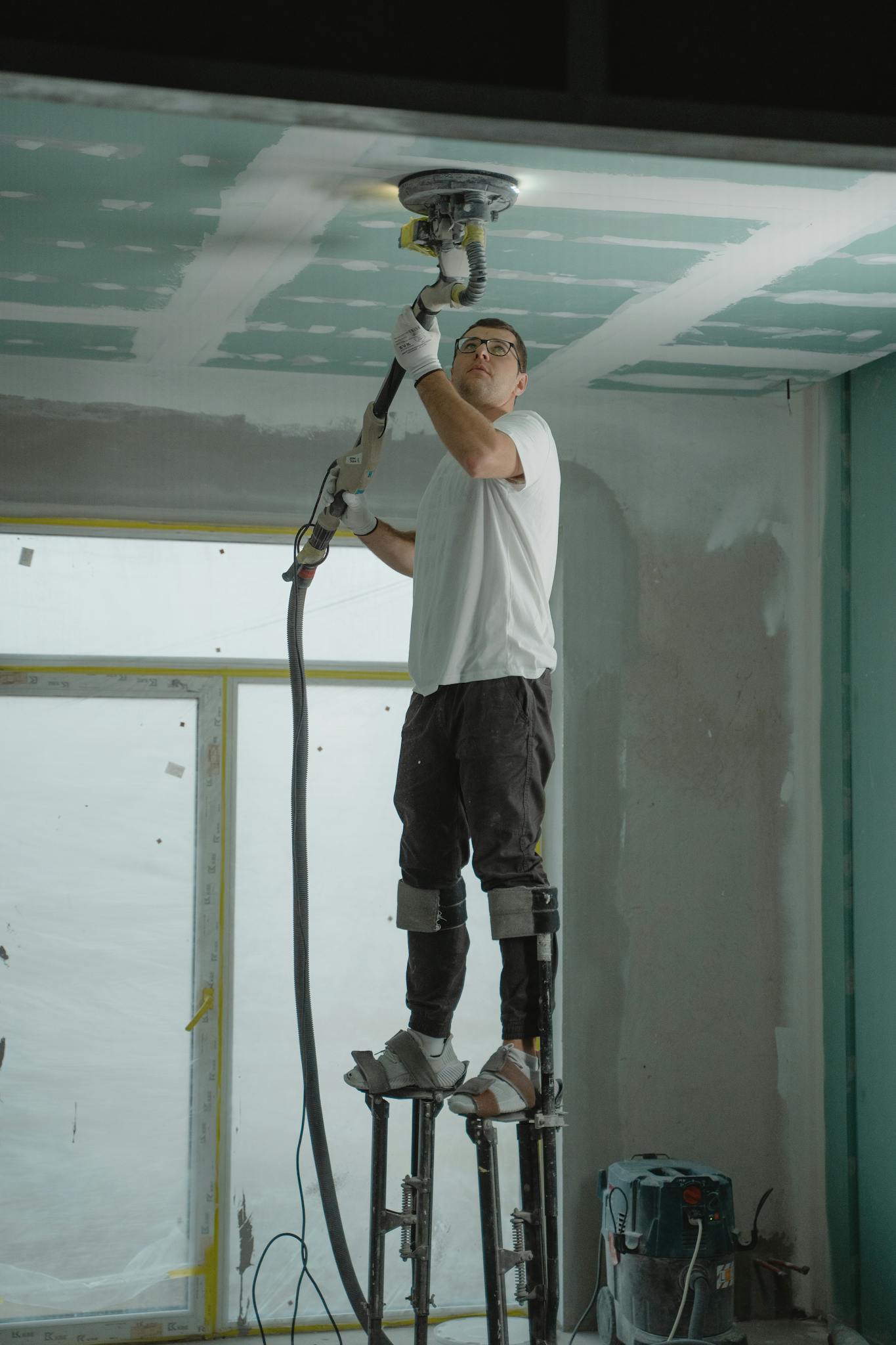 A man on stilts uses a ceiling sander for home renovation, focusing on drywall preparation.